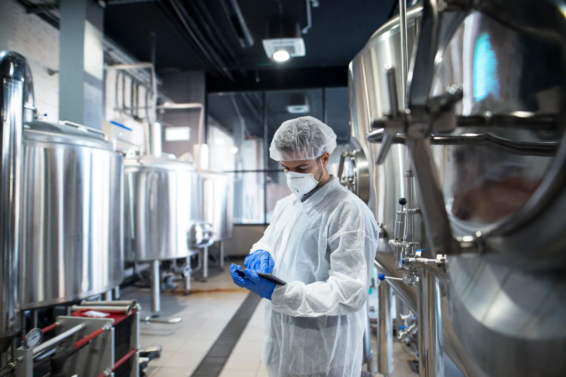 Technician in a hairnet, mask and white coverall checking a tablet beside stainless steel process tanks