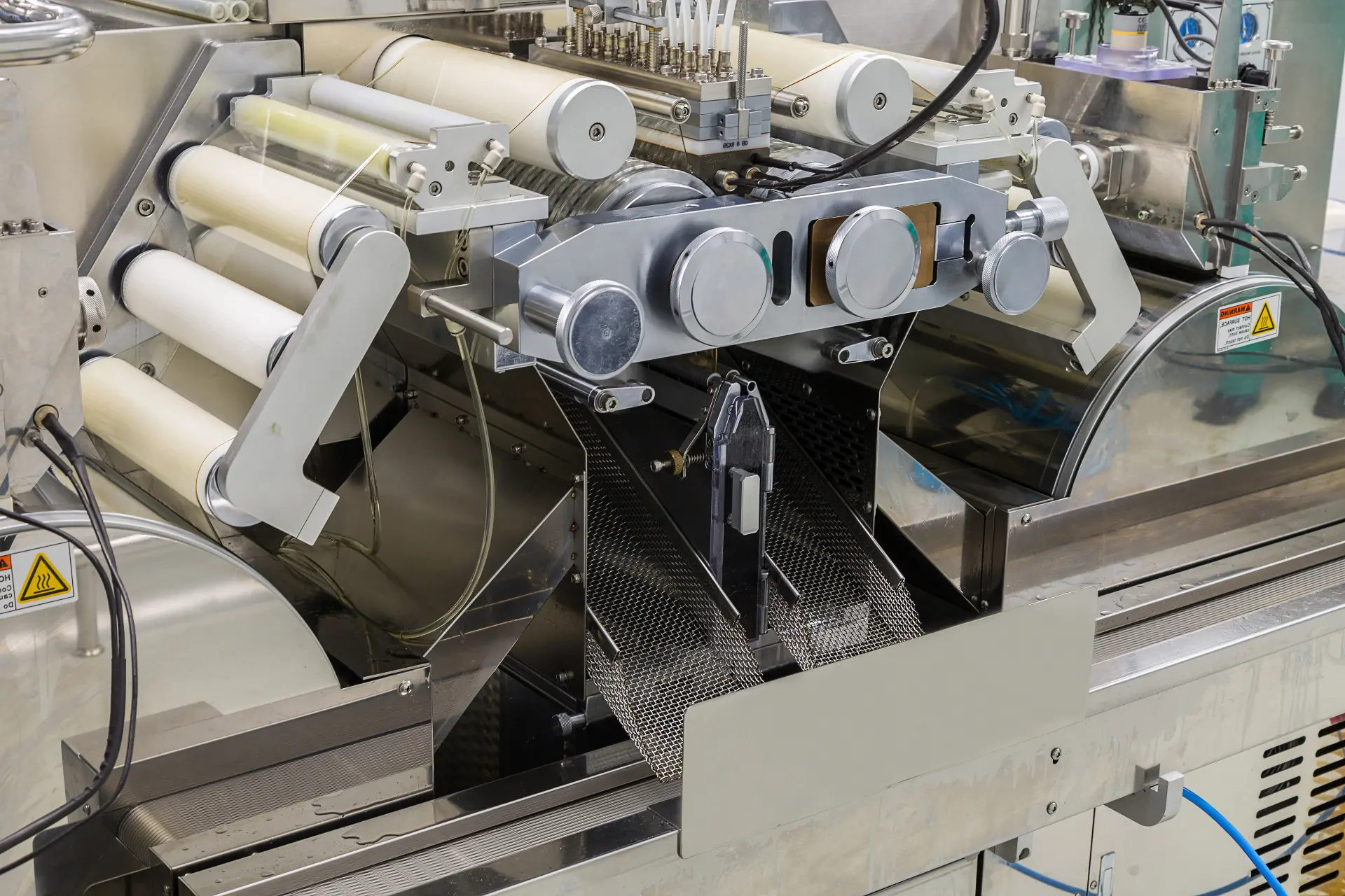 Close-up of a stainless steel industrial packaging machine with film rollers, guide arms and a mesh chute