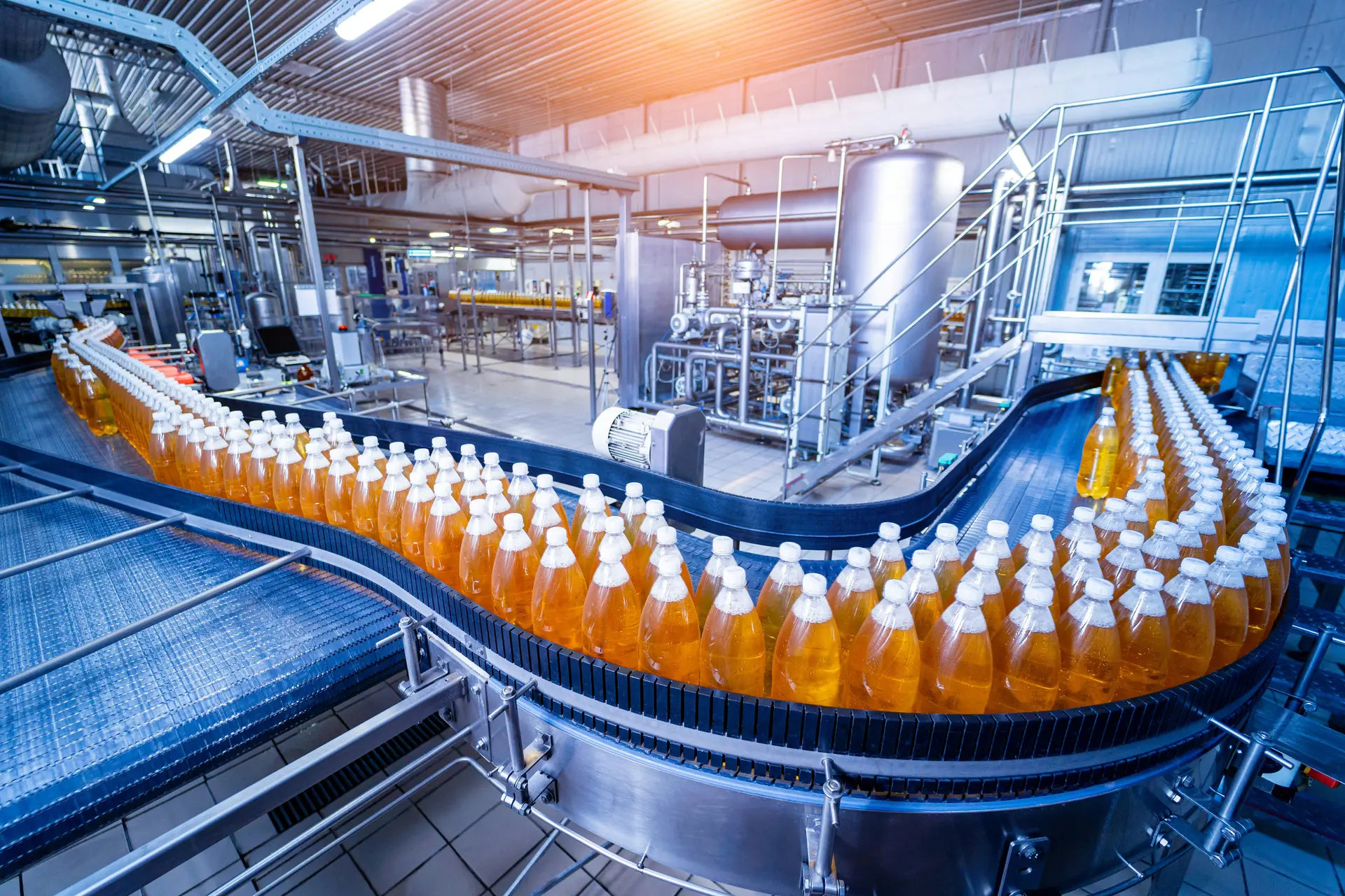 Curved conveyor system carrying rows of filled orange drink bottles through a busy bottling plant