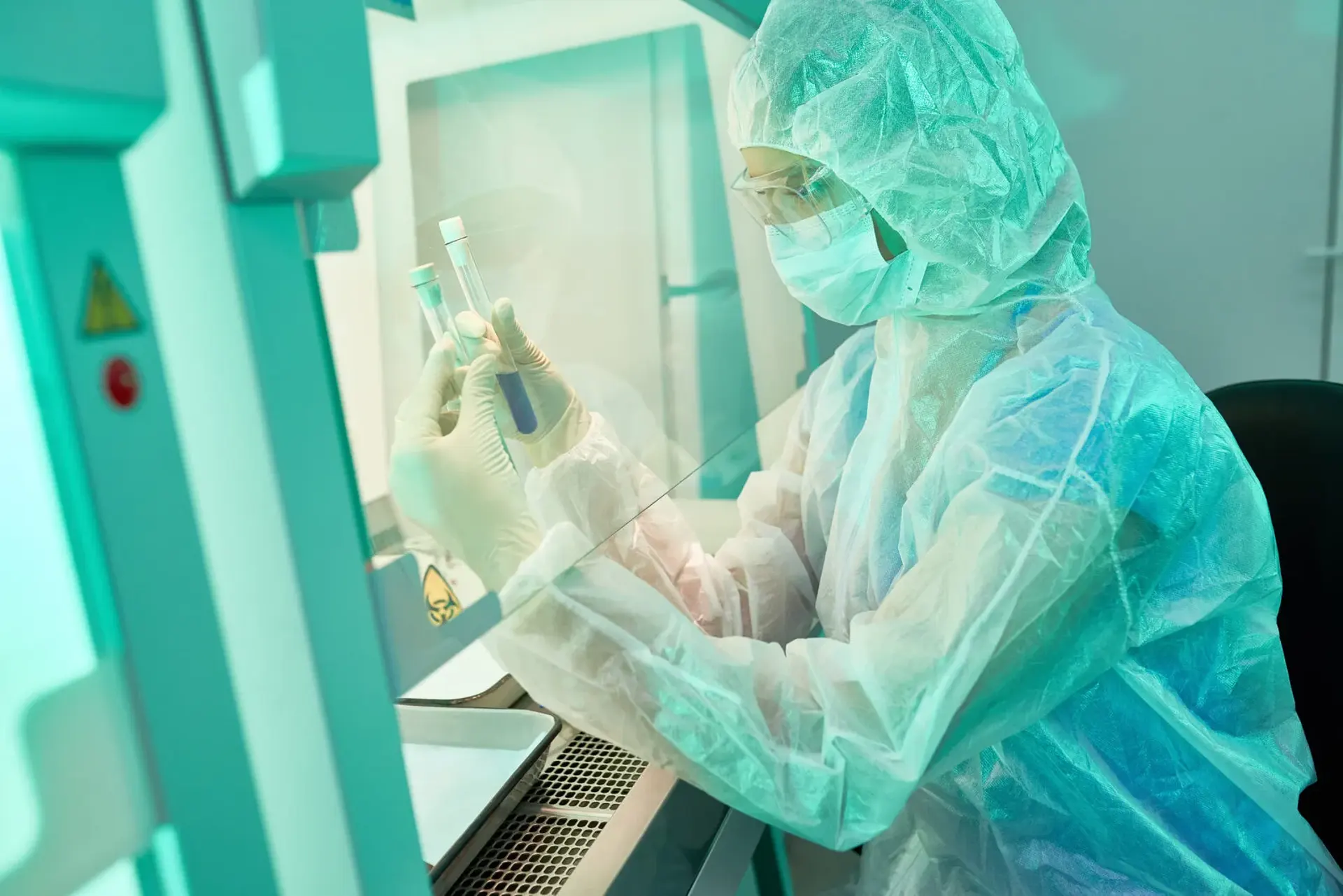 Technician in a protective suit and mask holding sample tubes at a laboratory safety cabinet with a glass sash