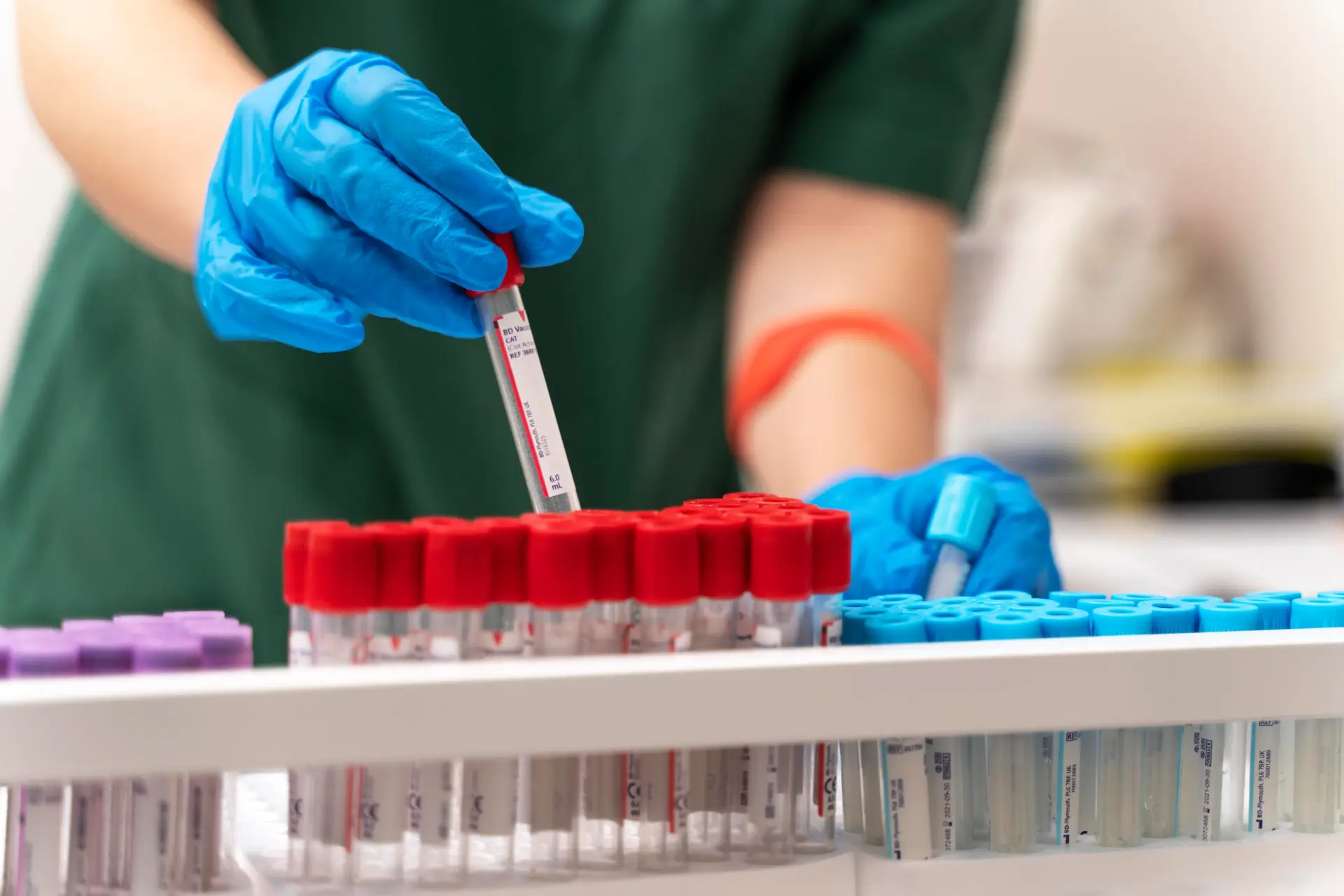 Gloved hand lifting a blood collection tube from a rack of red, blue and purple capped sample tubes