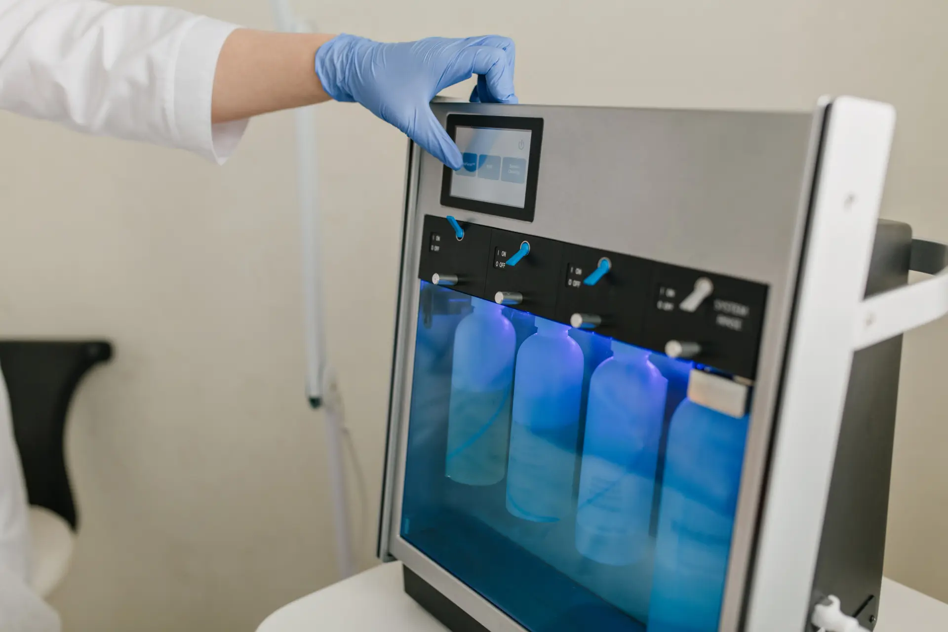Gloved hand pressing the touchscreen of a lab dispensing unit holding four bottles behind a blue-lit panel