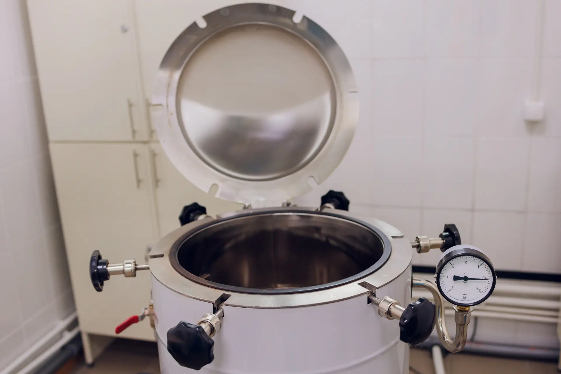 Stainless steel pressure vessel with its lid open, clamp handles and a pressure gauge in a tiled room