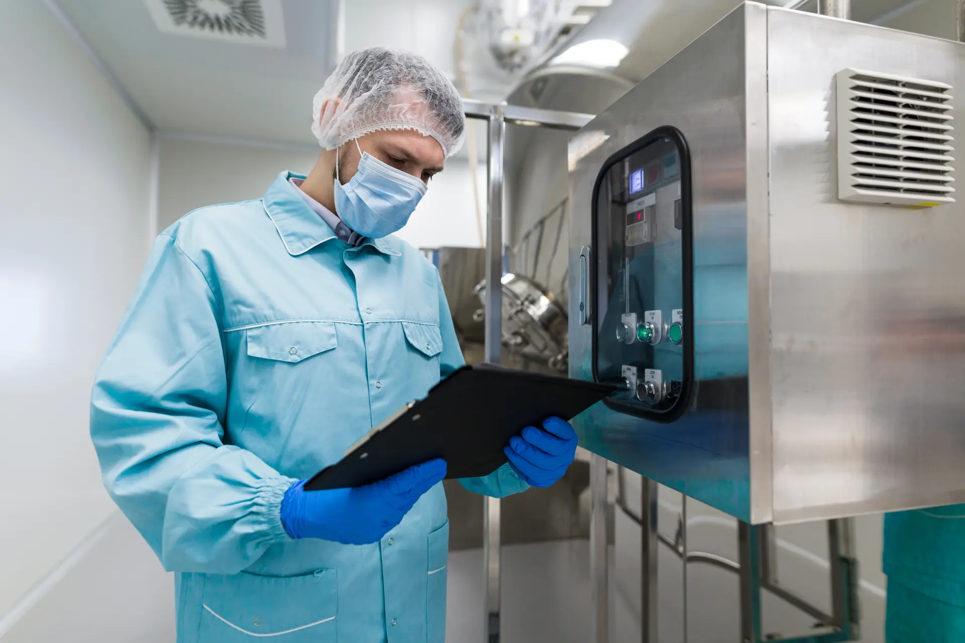 Technician in a cleanroom gown, hairnet and mask checking a clipboard beside a stainless steel control cabinet