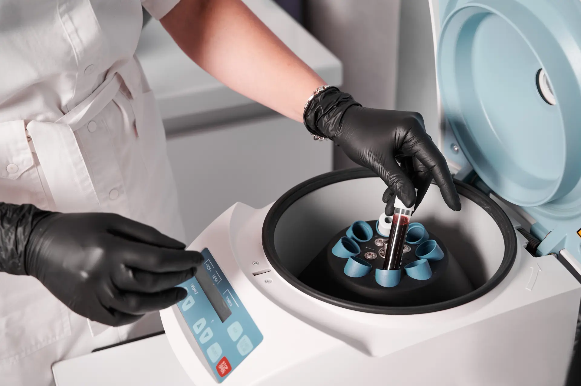 Gloved hands loading a blood sample tube into the rotor of an open benchtop centrifuge