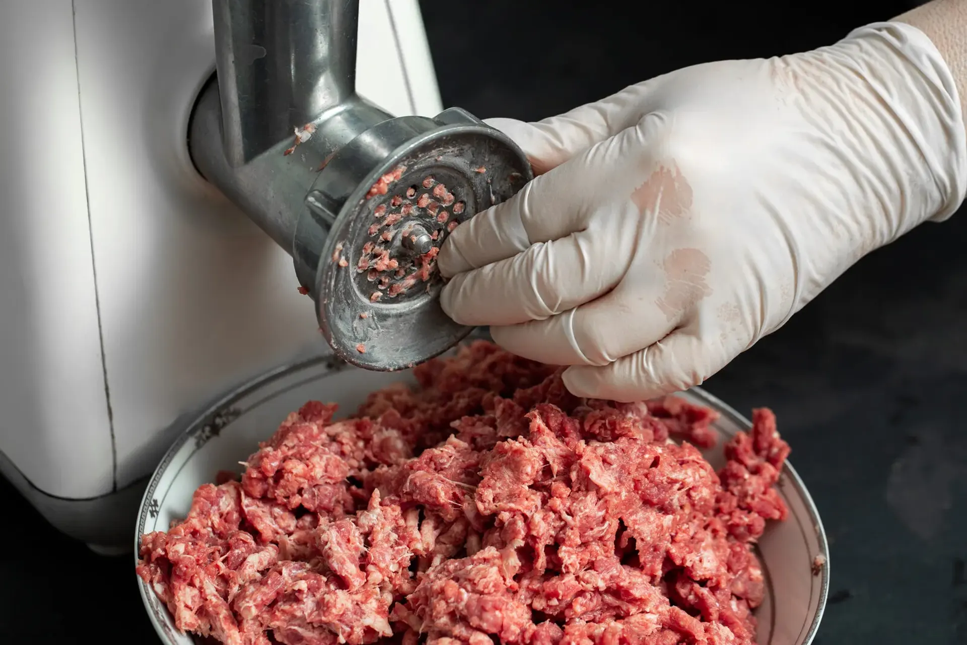 Gloved hand holding the head of a stainless steel meat grinder over a bowl of freshly minced meat