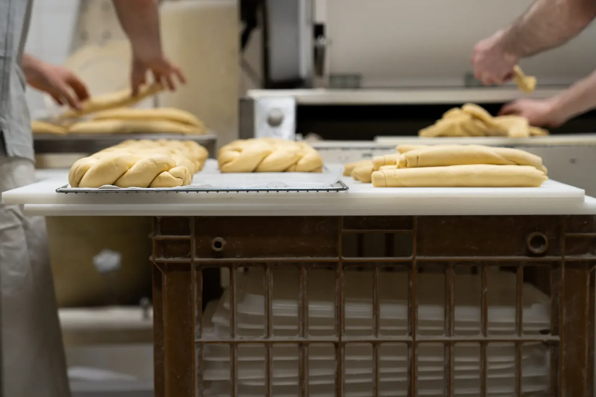 Portioned dough pieces and braided loaves on a bakery bench while bakers shape more dough in the background