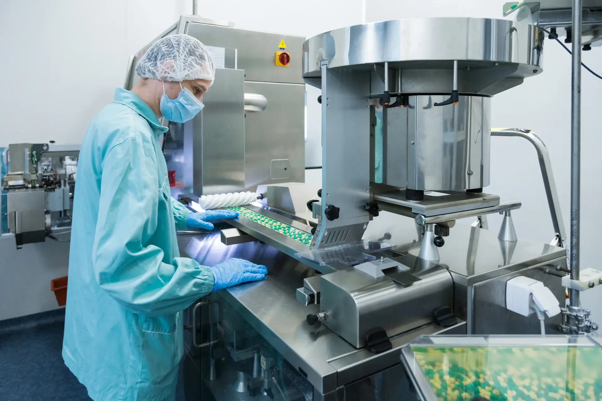 Technician in a cleanroom gown, hairnet and mask operating a pharmaceutical tablet packaging machine