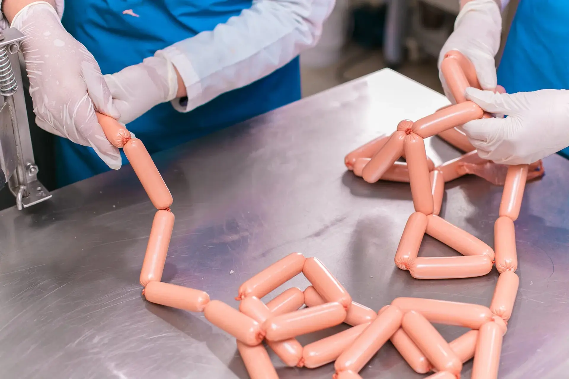 Workers in gloves and blue aprons arranging a chain of fresh sausages on a stainless steel table