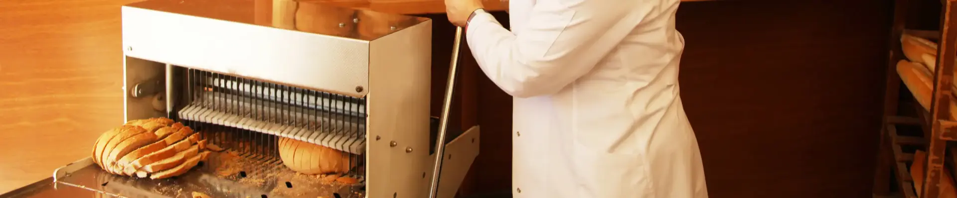 Baker in a white coat using a commercial bread slicer, with freshly sliced loaves on the machine tray