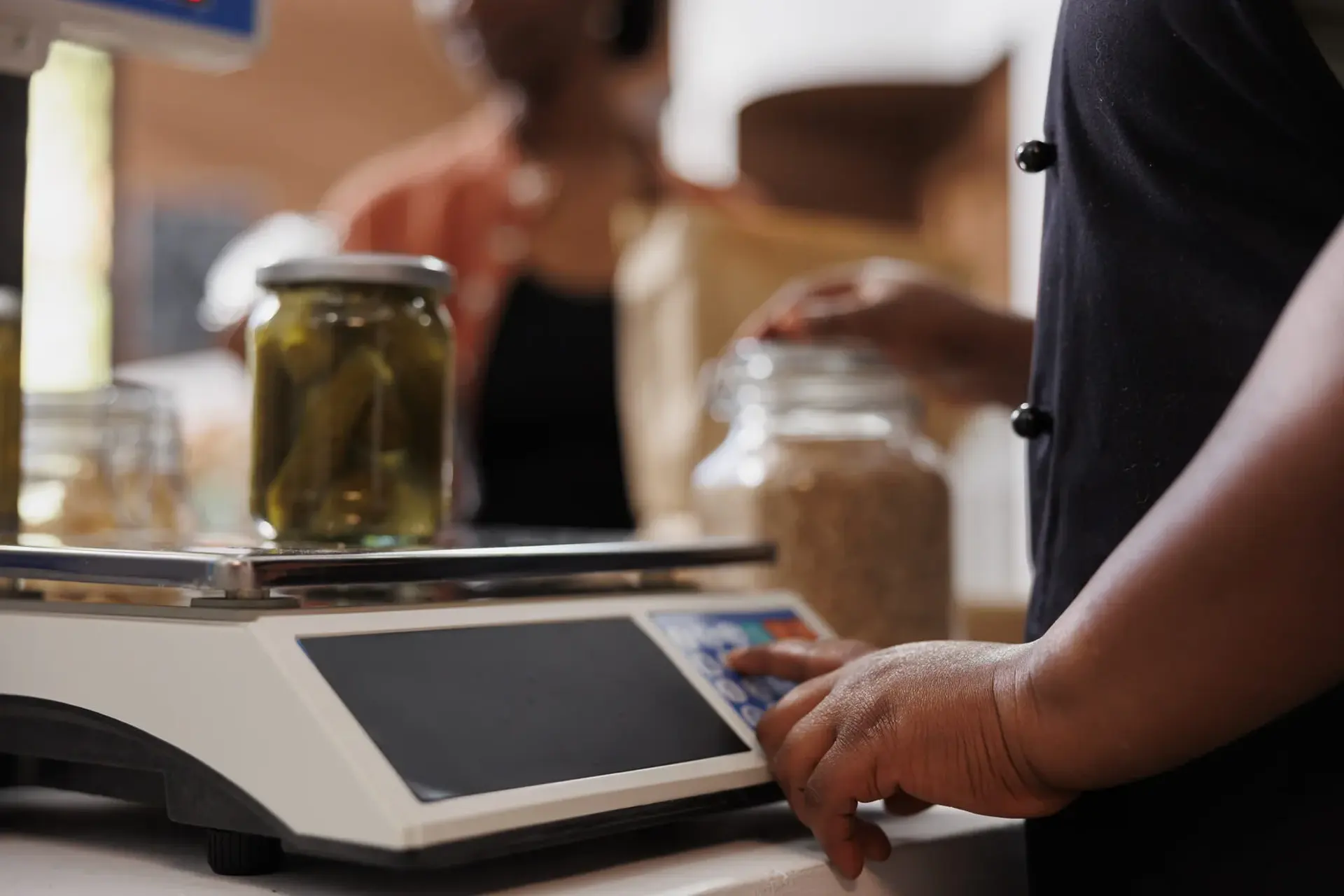 Glass jar of pickles sitting on a retail weighing scale while a person presses the keypad