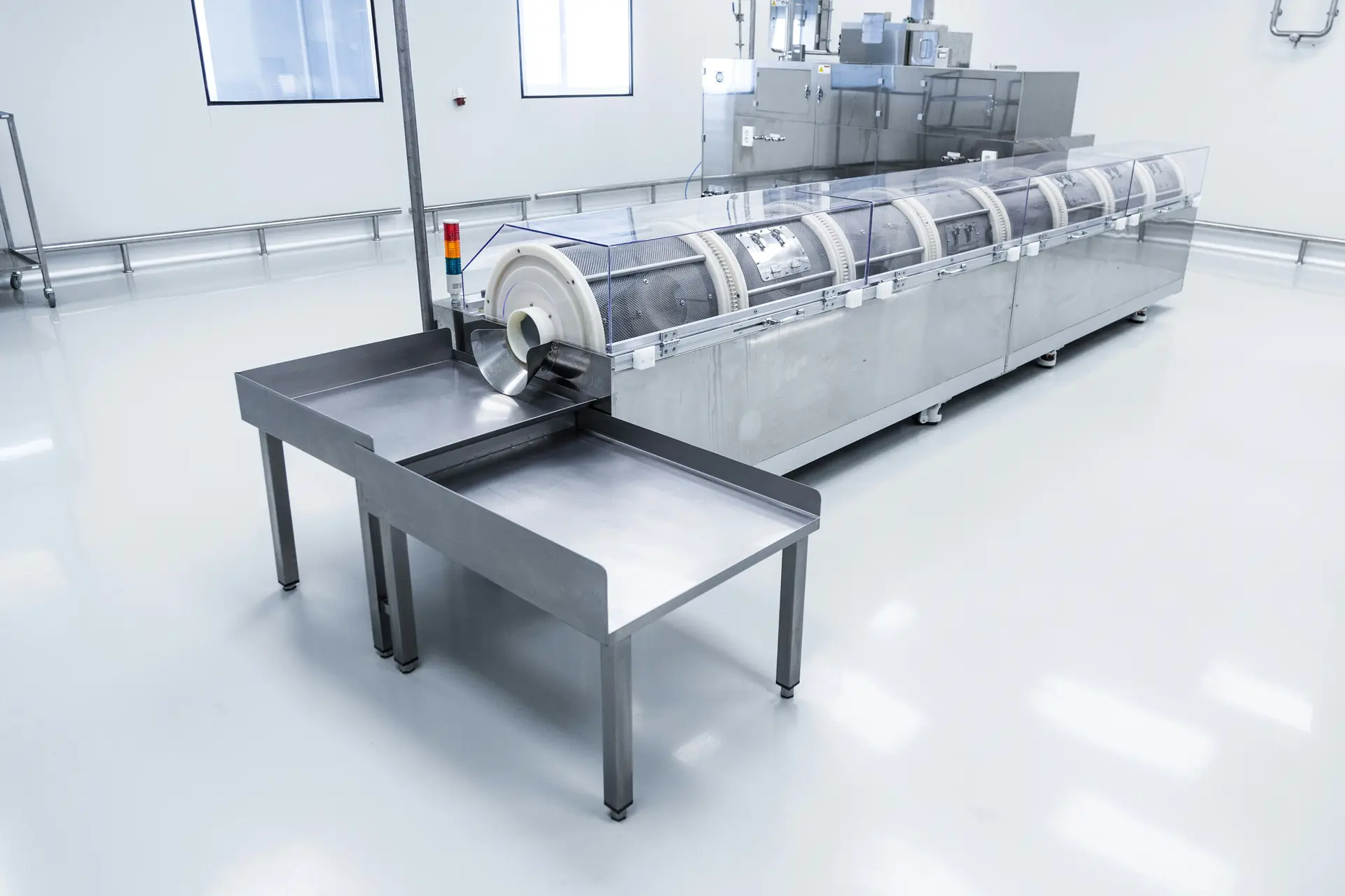 Long stainless steel drum processing machine with clear covers and a steel outfeed table in a white cleanroom