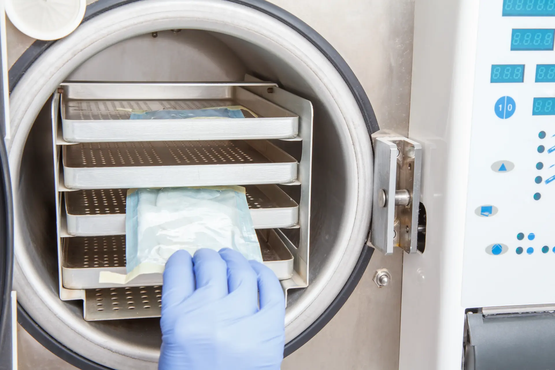 Gloved hand loading a sealed pouch onto the trays of an open chamber steriliser with a digital control panel