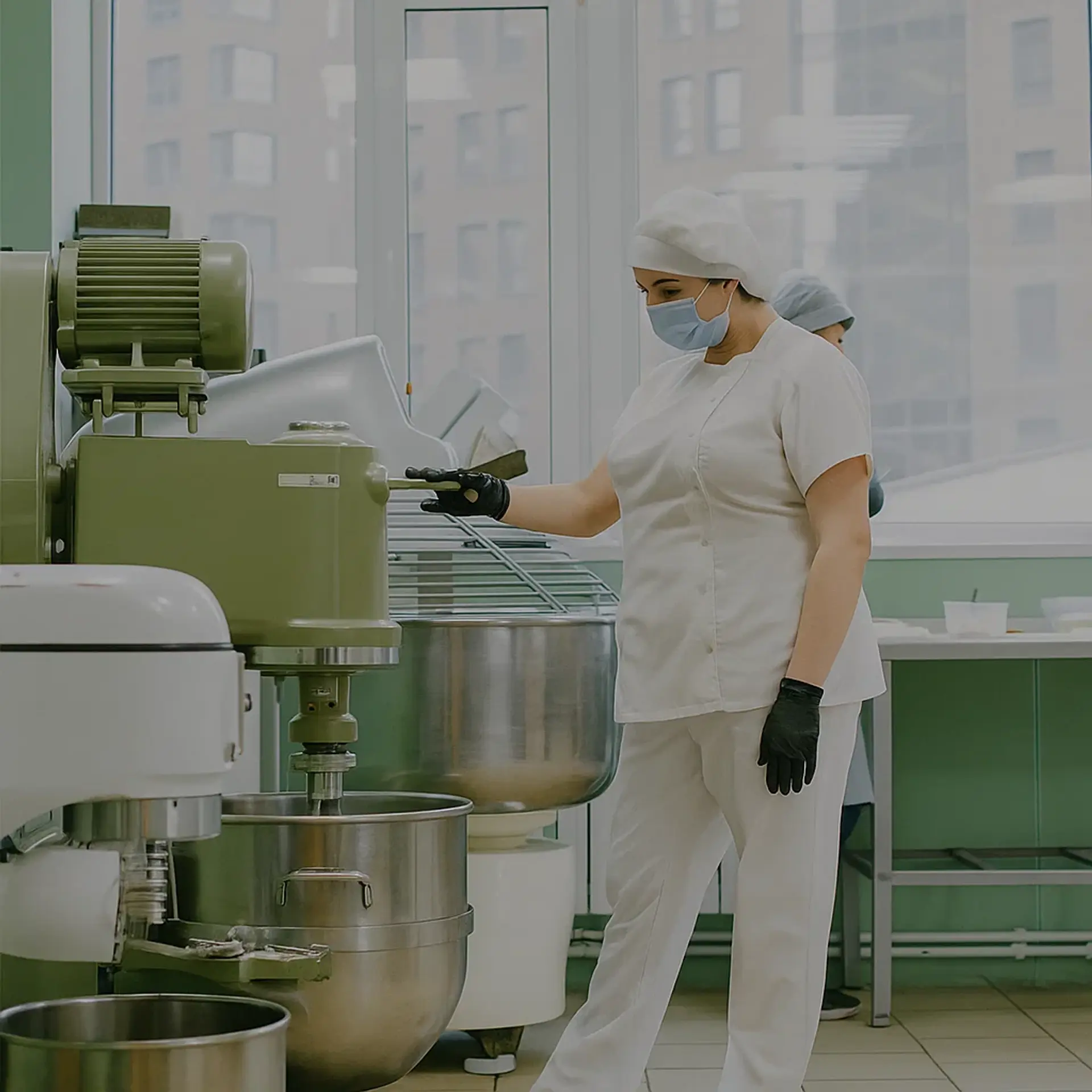 Baker in a hairnet, mask and gloves operating a large planetary mixer with stainless steel bowls in a bakery